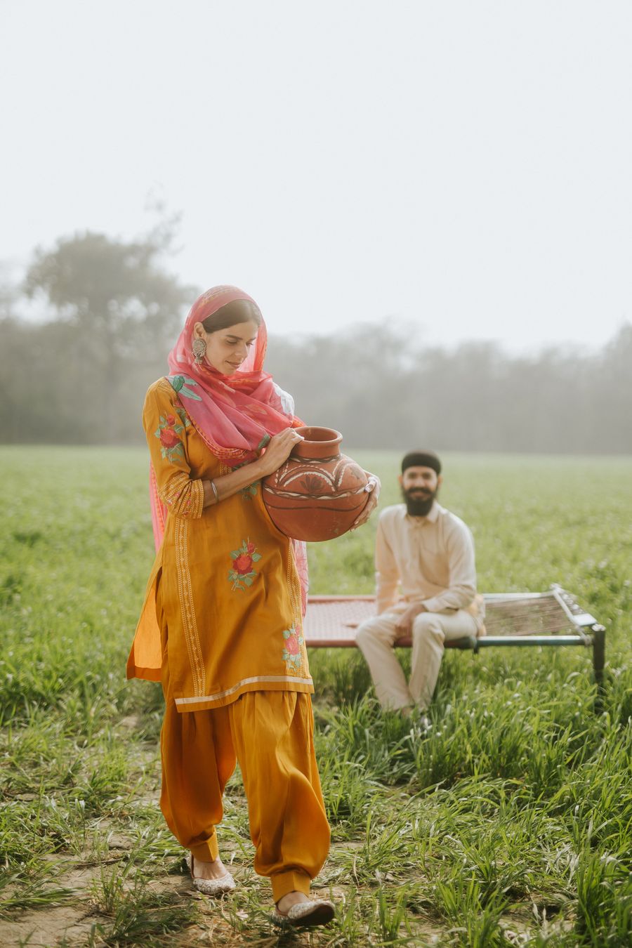 Naaz carrying a clay pot through the wheat fields, Himmat on the manji behind her